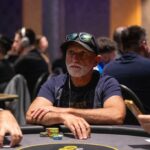 Ein Mann spielt Poker an einem Tisch mit grüner Filzdecke und hält eine Hand aus Spielkarten. Im Hintergrund sind Pokerchips und Getränke zu sehen.