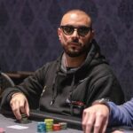 Ein Mann sitzt an einem Pokertisch und hält zwei Karten in der Hand. Er trägt eine dunkle Sonnenbrille und hat einen ernsten Ausdruck im Gesicht. Im Hintergrund sind weitere Spieler zu sehen