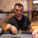 Ein Mann und eine Frau sitzen an einem Pokertisch und spielen Karten. Der Mann hat eine Hand mit Karten vor sich liegen, während die Frau ihre Karten hält. Im Hintergrund sind Pokerchips und Getr
