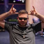 Ein Mann in einem dunkelblauen Anzug und einer roten Krawatte spielt konzentriert Poker an einem grünen Filztisch in einem eleganten Casino. Seine Hand hält Karten und Jetons liegen