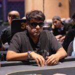 Ein Mann in elegantem Anzug sitzt an einem Pokertisch und hält zwei Spielkarten in der Hand. Er wirkt konzentriert und fokussiert auf das Spiel. Im Hintergrund sind Pokerchips und