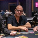 Ein Mann und eine Frau spielen Poker an einem eleganten Tisch in einem stilvollen Raum mit Kronleuchterbeleuchtung. Der Mann hat Karten in der Hand und die Frau betrachtet ihre Karten nachdenklich