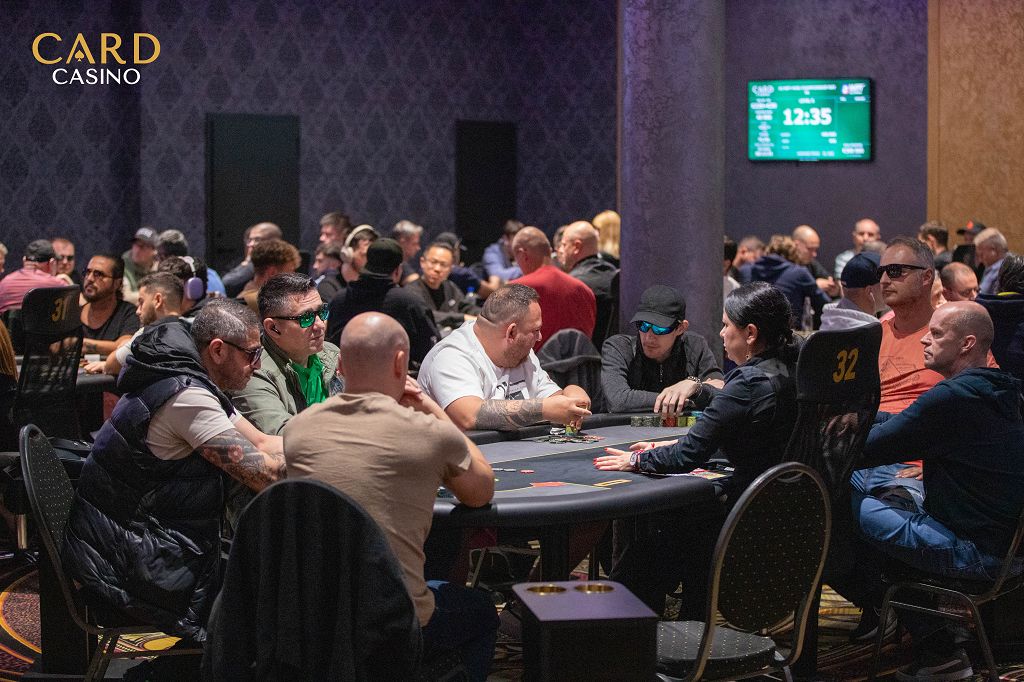 Ein Mann in einem dunklen Anzug und einer roten Krawatte sitzt an einem Pokertisch und hält seine Karten in der Hand, während er konzentriert auf den Tisch schaut. Im Hintergrund sind