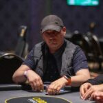 Ein Mann sitzt an einem Pokertisch, hält Karten in der Hand und schaut konzentriert auf den Tisch. Andere Spieler sind im Hintergrund zu sehen.