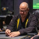 Ein Mann sitzt an einem Pokertisch und hält zwei Karten in der Hand. Er hat eine ernste Miene und starrt konzentriert auf den Tisch. Andere Spieler sind im Hintergrund verschwommen