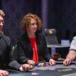 Ein junger Mann sitzt an einem Pokertisch und betrachtet konzentriert seine Karten. Im Hintergrund sind andere Spieler und Pokerchips zu sehen.