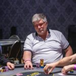 Ein Mann sitzt an einem Pokertisch und hält seine Karten in der Hand, während er konzentriert auf den Tisch schaut. Andere Spieler und Chips sind im Hintergrund zu sehen.