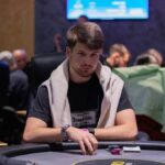 Ein Mann und eine Frau sitzen an einem Pokertisch und halten ihre Karten. Sie haben konzentrierte Gesichtsausdrücke. Im Hintergrund sind Pokerchips und Getränke zu sehen.