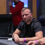 Ein Mann sitzt an einem Pokertisch und hält zwei Spielkarten in der Hand. Er scheint konzentriert zu sein, während er auf seine Gegner blickt. Die Karten und Chips auf dem Tisch de