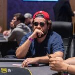 Ein Mann und eine Frau sitzen an einem Pokertisch und spielen Karten. Der Mann hat eine Hand mit Chips vor sich liegen, während die Frau konzentriert ihre Karten betrachtet. Die Szene spielt in einem