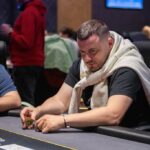 Ein Mann und eine Frau spielen Poker an einem eleganten Tisch in einem stilvollen Raum mit Kronleuchter. Der Mann hat eine Hand mit Karten, die er prüft, während die Frau konzentriert ihre eigenen K