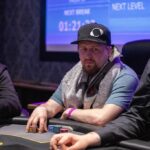 Ein glücklicher Mann sitzt an einem Pokertisch und hält zwei Karten in der Hand, während er lächelnd auf seine Gegner schaut. Die Spielszene ist von Chips und Spielkarten umgeben.