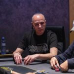 Ein professioneller Pokertisch mit grünem Filz, umgeben von Spielern, die Karten halten und Pokerchips stapeln. In der Mitte des Tisches liegen Spielkarten und Jetons. Im Hintergrund