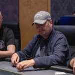 Ein Mann sitzt an einem Pokertisch und hält zwei Karten in der Hand. Er hat eine Pokerface und schaut konzentriert auf den Tisch mit Chips und Karten. Im Hintergrund sind andere Spieler zu sehen
