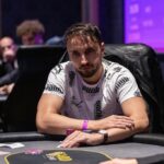 Ein Mann in einem eleganten dunkelblauen Anzug sitzt an einem Pokertisch und hält zwei Karten in der Hand, während er konzentriert auf seine Gegner blickt. Die Szene ist in einem stil