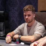 Ein Mann in einem dunklen Anzug sitzt an einem Pokertisch und hält eine Hand mit Spielkarten. Er schaut konzentriert auf seine Karten, während im Hintergrund weitere Spieler und Chips zu sehen sind.