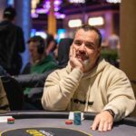 Ein Mann in einem schwarzen Anzug und einer Sonnenbrille sitzt an einem Pokertisch und hält zwei Spielkarten in der Hand. In der Mitte des Tisches liegen weitere Karten und Pokerchips.