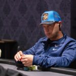 Ein Mann sitzt an einem Pokertisch und hält seine Karten in der Hand, während er konzentriert auf die Karten auf dem Tisch schaut. Andere Spieler sind im Hintergrund zu sehen, die auf ihre