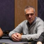 Ein professioneller Pokertisch mit grünem Filz und einem Stapel von Pokerchips in verschiedenen Farben, umgeben von Spielkarten und Getränken. Vier Personen sind um den Tisch herum versammelt