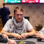 Ein Mann sitzt an einem Pokertisch und hält zwei Karten in der Hand, während er konzentriert auf die Gemeinschaftskarten in der Mitte des Tisches schaut. Andere Spieler sind im Hintergrund