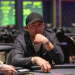 Ein Mann in einem eleganten Anzug und einer schwarzen Krawatte sitzt an einem Pokertisch und hält ein Ass und eine Pik-Dame in der Hand. Die anderen Spieler am Tisch haben ernste Gesichtsausdr
