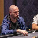 Ein Mann und eine Frau sitzen lächelnd an einem Pokertisch, während sie Karten halten und auf ihre Gegner schauen. Der Mann hat eine Sonnenbrille auf und die Frau trägt eine elegante Abendgard