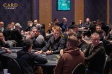 Ein Mann sitzt an einem Pokertisch und hält seine Karten in der Hand, während er konzentriert auf den Tisch schaut. Um ihn herum sind Pokerchips und Getränke zu sehen.