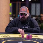 Ein Mann spielt Poker und hält eine Hand mit Karten. Er trägt einen grauen Anzug und sitzt an einem Pokertisch in einem eleganten Casino.