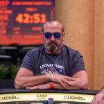Ein glücklicher Mann sitzt an einem Pokertisch und hält eine Hand mit Spielkarten. Er lächelt in die Kamera und wirkt entspannt. Im Hintergrund sind weitere Spieler und Pokerchips zu sehen