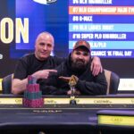 Ein Mann sitzt an einem Pokertisch und hält seine Karten in der Hand, während er konzentriert auf den Tisch schaut. Im Hintergrund sind andere Spieler und Chips zu sehen.