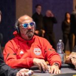Ein Mann sitzt an einem Pokertisch und hält zwei Spielkarten in der Hand. Neben ihm stehen Pokerchips und Getränke. Im Hintergrund sind weitere Spieler und eine beleuchtete Bar zu sehen.