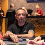 Ein grüner Pokertisch mit Spielkarten, Pokerchips und Getränken darauf, umgeben von Personen in einer entspannten Atmosphäre.