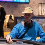 Ein Tisch mit grünem Filz, auf dem Pokerkarten und Pokerchips liegen, umgeben von vier Personen, die konzentriert Poker spielen.