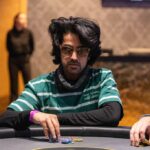 Ein Mann sitzt an einem Pokertisch und hält zwei Karten in der Hand, während er konzentriert auf die Gemeinschaftskarten auf dem Tisch schaut. Andere Spieler sind im Hintergrund zu sehen.