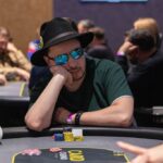 Ein Mann mit einem roten Hut und einer dunklen Brille spielt Poker an einem grünen Filztisch mit Chips und Karten vor sich.