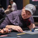 Ein Mann und eine Frau sitzen an einem Pokertisch und halten Karten. Sie schauen konzentriert auf ihre Hände, während im Hintergrund Casino-Chips und Getränke zu sehen sind.