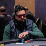 Ein Mann und eine Frau sitzen an einem Pokertisch, während sie konzentriert ihre Karten betrachten. Die Frau hat blonde Haare und trägt eine schwarze Bluse, der Mann trägt ein weißes Hemd.