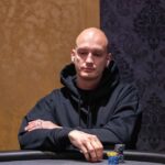 Ein Mann in einem dunkelblauen Anzug und einer roten Krawatte sitzt an einem Pokertisch und hält zwei Spielkarten in der Hand. Er hat einen konzentrierten Blick und spielt Poker.