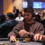 Ein Bild von fünf Personen, die an einem Pokertisch sitzen und Karten spielen. Es herrscht eine konzentrierte Atmosphäre, während sie ihre Karten betrachten.