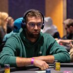 Ein Bild von fünf Personen, die um einen Pokertisch sitzen und Karten spielen. Es herrscht eine konzentrierte Atmosphäre, während sie ihre Karten betrachten. Es gibt Pokerchips und Geträn