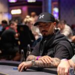 Ein Mann in schwarzer Kleidung und Sonnenbrille spielt Poker an einem Tisch mit Chips und Karten. Er hat eine ernste Miene und konzentriert sich auf sein Blatt.