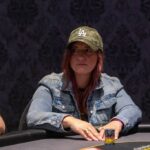 Ein Mann sitzt an einem Pokertisch und hält zwei Spielkarten in der Hand, während er ernsthaft auf seine Gegner blickt. Die Atmosphäre ist konzentriert und spannungsgeladen.