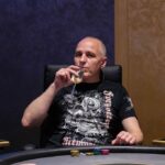 Ein Mann sitzt an einem Pokertisch und hält zwei Spielkarten in der Hand, während er konzentriert auf die Karten auf dem Tisch schaut. Andere Spieler sind im Hintergrund zu sehen.