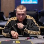 Ein professioneller Pokerspieler mit Sonnenbrille und Hut sitzt an einem Pokertisch mit Chips und Karten vor sich.