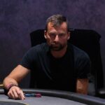 Ein Mann und eine Frau sitzen an einem Pokertisch und spielen eine Partie Poker. Der Mann hat eine Hand mit Karten in der Hand und schaut konzentriert auf den Tisch, während die Frau ihm gegenüber mit ern