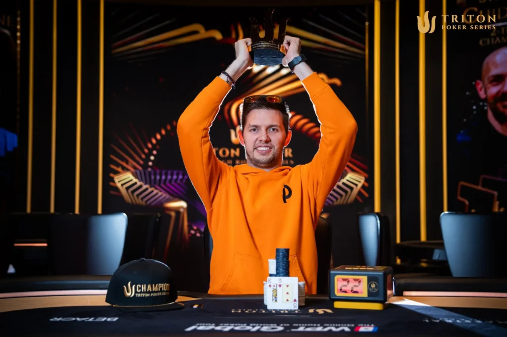 Ein Mann sitzt an einem Pokertisch in einem Casino. Er trägt eine schwarze Kappe und konzentriert sich auf seine Karten. Im Hintergrund sind weitere Spieler und Dealer zu sehen.