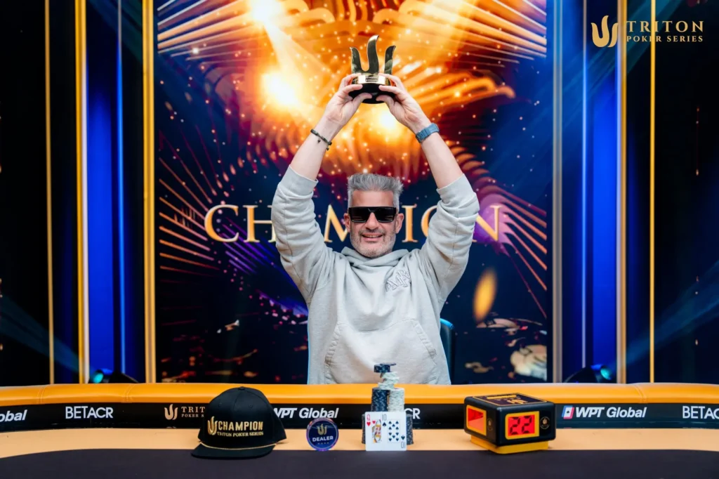 Ein Mann in einem schwarzen T-Shirt und einer Baseballkappe sitzt an einem Pokertisch und betrachtet konzentriert seine Karten. Im Hintergrund sind andere Spieler und Pokerchips zu sehen.