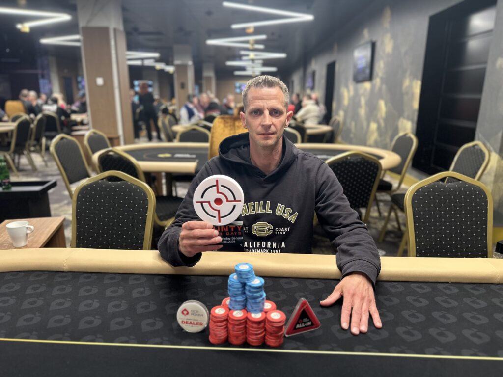 Ein Pokertisch mit Spielkarten, Jetons und einem Dealer-Button. Spieler sitzen um den Tisch herum und warten gespannt auf ihre Karten. Es herrscht eine konzentrierte Atmosphäre.