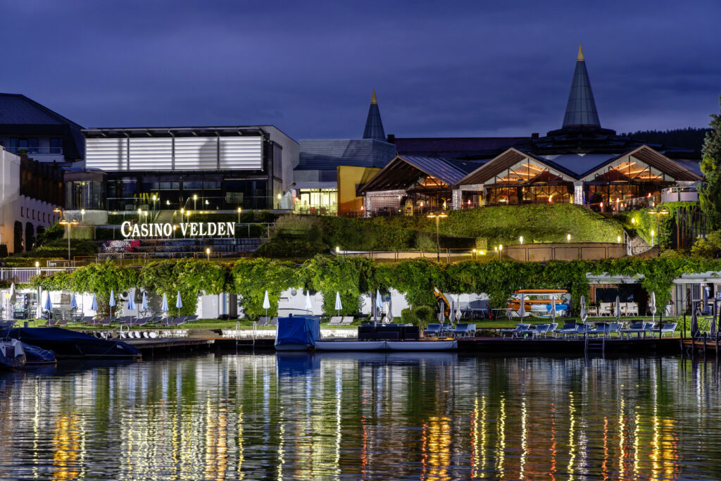 Ein nächtliches Bild von Velden am Wörthersee. Die beleuchtete Promenade spiegelt sich im ruhigen Wasser des Sees wider, während im Hintergrund die Lichter der Stadt funkeln.