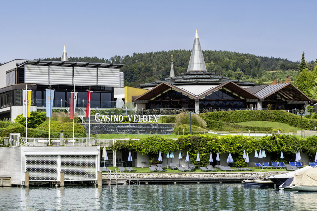 Ein sonniger Tag in Velden am Wörthersee: Ein elegantes Gebäude mit blumengeschmückten Balkonen, umgeben von grünen Bäumen und einem strahlend blauen Himmel.